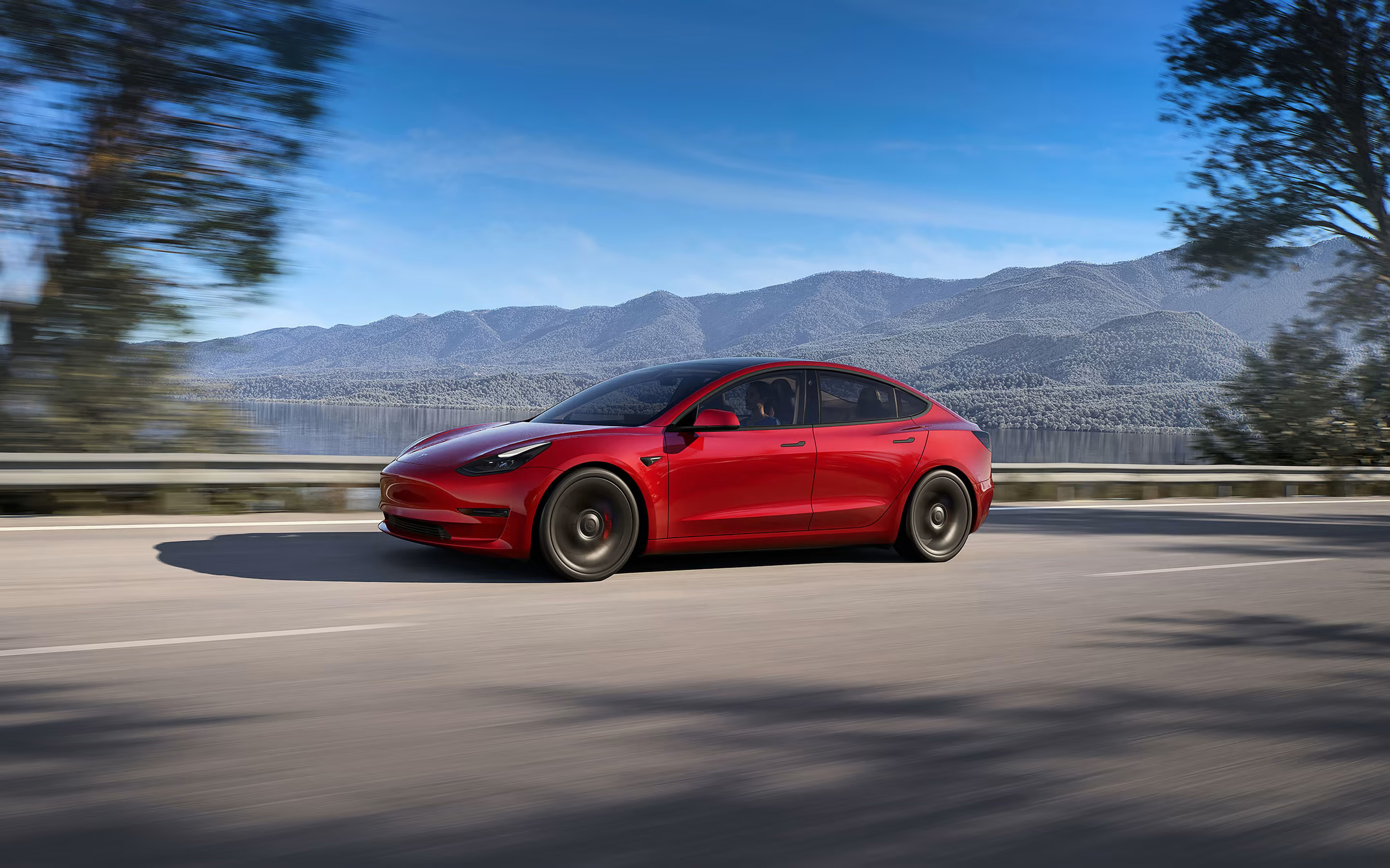 Red Model 3 driving down a lakeside road